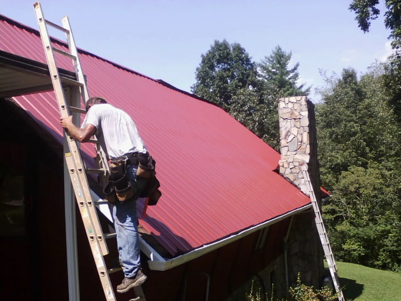 Roofer on a ladder beside a red metal roof and stone chimney — Soffit & Fascia Repair by Roofing Brothers in Woodlake